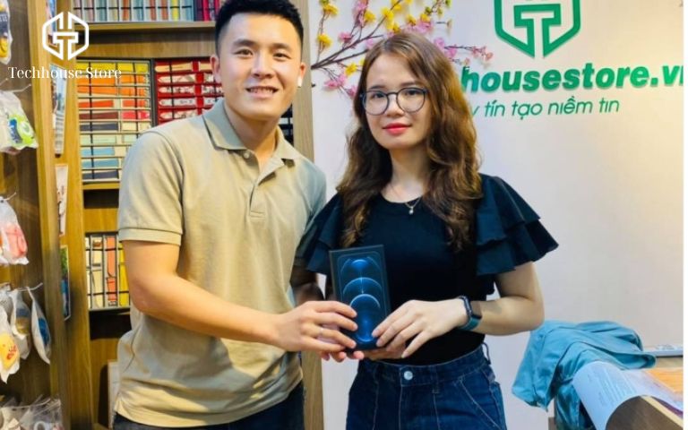 Cách kiểm tra iPhone cũ zin 100% trước khi mua (Chi tiết A-Z) Nhân viên Tech House Store đang trao hộp máy iPhone 12 Pro Max cho khách hàng nữ đeo kính tại cửa hàng.