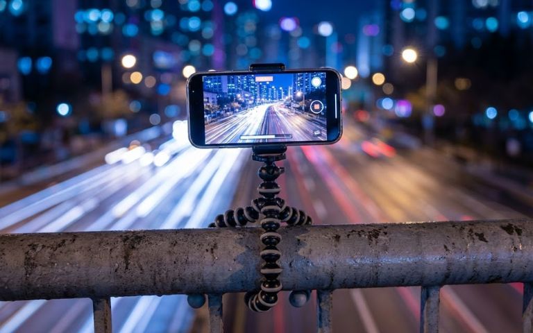 Top 15+ App Chụp Ảnh Đẹp Trên iPhone Miễn Phí 2026 iPhone được gắn trên tripod mini đang thực hiện phơi sáng (long exposure) cảnh thành phố về đêm với các vệt đèn xe.