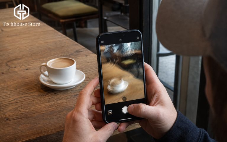 Góc nhìn qua vai người dùng đang cầm iPhone chụp ảnh một tách cà phê, cảnh bên ngoài rõ nét nhưng hình ảnh trên màn hình điện thoại bị nhòe và gợn sóng do lỗi camera.