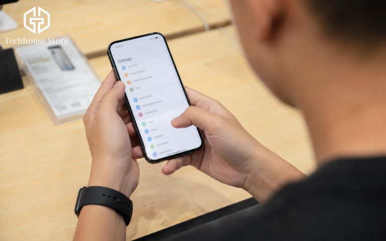Mua iPhone cũ trả góp tại Hà Nội: cần chuẩn bị những gì? Chụp qua vai (over-the-shoulder) một người đang cầm điện thoại bằng hai tay, ngón tay cái đang vuốt màn hình hiển thị một giao diện cài đặt chung chung để kiểm tra chức năng.