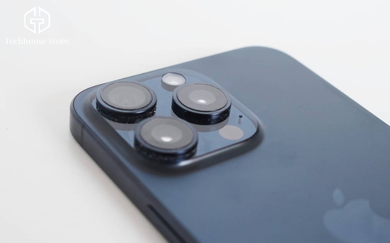 Nên chọn iPhone 128GB hay 256GB? Cách chọn dung lượng đúng theo nhu cầu Chụp cận cảnh cụm ba camera lớn mặt lưng thiết bị điện thoại cao cấp bằng kim loại nhám, đặt trên mặt bàn gỗ với ánh sáng tự nhiên hắt vào.