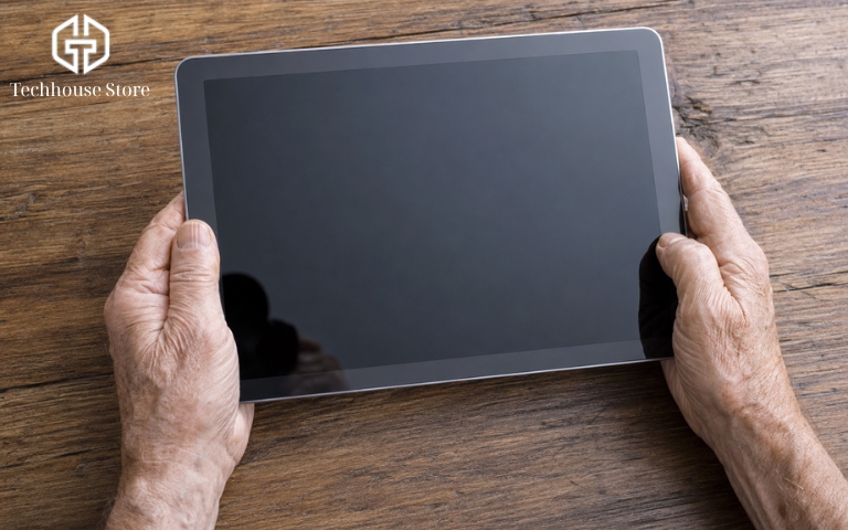 iPad cho người lớn tuổi: Cách chọn máy phù hợp, dễ sử dụng Cận cảnh hai bàn tay có nếp nhăn của người lớn tuổi đang cầm một chiếc máy tính bảng màu xám trên nền một chiếc bàn gỗ, thể hiện rõ tỷ lệ kích thước giữa máy và tay.