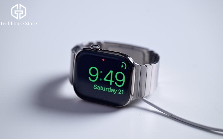 Cách sạc Apple Watch đúng cách cho người mới sử dụng Apple Watch Series 10 với dây kim loại đặt trên bộ sạc, màn hình hiển thị đồng hồ số màu xanh và biểu tượng nhận sạc.