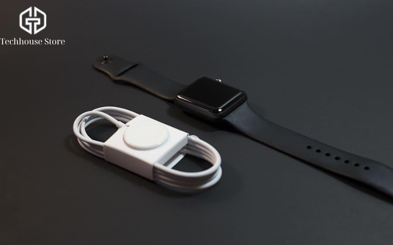 Cách sạc Apple Watch đúng cách cho người mới sử dụng Đồng hồ Apple Watch màu đen đặt cạnh dây sạc từ tính màu trắng trên nền tối, minh họa phụ kiện cần chuẩn bị khi sạc.